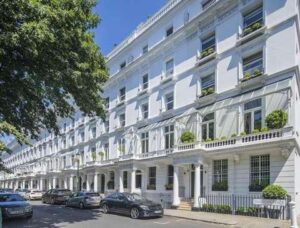 A row of elegant white terraced houses with tall windows and ornate balconies on a sunny day. Some houses have potted plants on their balconies. Several cars are parked along the street, and a large tree provides shade on the left—truly a Knightbridge development opportunity by Remi Charles.