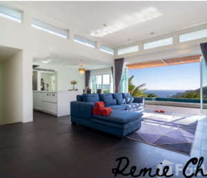 A modern, open-concept living room with a blue sectional sofa facing a large sliding glass door that opens to a scenic ocean view. The room has white walls, high windows, and a sleek kitchen area in the background. A chandelier hangs in the dining area.