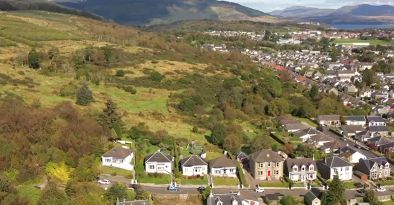A panoramic view of a picturesque town set against a backdrop of rolling green hills and mountains. The foreground shows rows of houses with sloping roofs and well-kept gardens, transitioning to lush, wooded hills. Picture of land in Dunoon for sale by auction with World of Auctions and Palace Auctions.