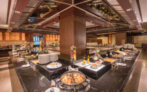 A lavish buffet setup at the Grand Pacific Sovereign Resort & Spa features a variety of dishes on display. Neatly arranged plates, bowls, trays of fruits, eggs, sauces, and a selection of hot and cold foods complement the room's modern decor with wood and glass elements and ample lighting.