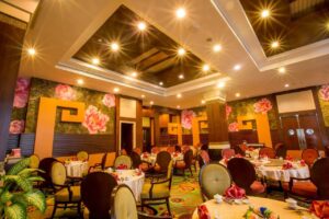 A warmly lit, elegant restaurant interior at the Grand Pacific Sovereign Resort & Spa features floral wallpaper, round tables covered with white cloths, and cushioned chairs. Each table is set with red napkins and dinnerware. Recessed and pendant lighting illuminate the room, creating a cozy ambiance.