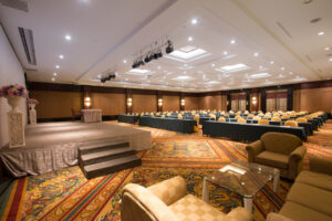 A spacious, well-lit conference room at the Grand Pacific Sovereign Resort & Spa features a carpeted floor, rows of tables with chairs facing a stage, and comfortable armchairs around glass tables at the front. The stage is equipped with a podium and decorated with a floral arrangement.