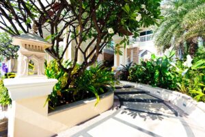 A sunlit garden path surrounded by lush greenery leads to a building with large white columns and balconies, reminiscent of the opulence found at the Grand Pacific Sovereign Resort & Spa. The path features geometric designs and is bordered by bushes, palms, and flowering trees. A decorative stone lantern sits beside the path on the left.