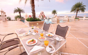 A luxurious outdoor dining area by the pool at the Grand Pacific Sovereign Resort & Spa, adorned with statues and palm trees. A table is set with plates of breakfast items, glasses of orange juice, and cups of coffee. The setting overlooks an ocean view with a clear sky during sunrise or sunset.