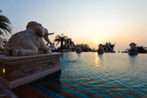 A serene outdoor pool at the Grand Pacific Sovereign Resort & Spa boasts water features shaped like elephants. The horizon displays a beautiful sunset, casting a golden glow across the water. Ornate buildings with pointed roofs are visible in the background, framed by palm trees—an exclusive off-market hotel investment opportunity.