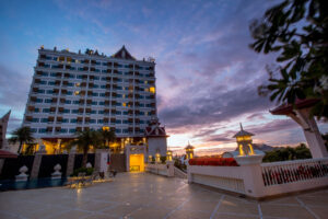 A tall hotel building with lit windows stands against a dramatic evening sky with hues of blue, purple, and pink. In the foreground, the spacious terrace of the Grand Pacific Sovereign Resort & Spa features outdoor seating and lantern-style lights. The scene exudes a serene and luxurious atmosphere.