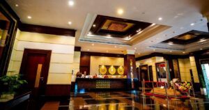 A spacious, well-lit hotel lobby with polished dark brown flooring, a front desk adorned with art, and a decorative sitting area with plush seating. Walls are cream-colored with wooden accents and there is a large mirror on the left. Just 180 kilometres from Bangkok, this serene locale offers breathtaking views across the Gulf of Thailand.