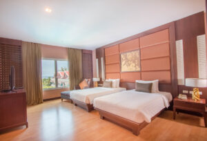 A spacious room at the Grand Pacific Sovereign Resort & Spa features two neatly made double beds, a large window with beige curtains offering a view of trees and rooftops, wooden flooring, a flat-screen TV on a dark wood dresser, and modern decor in brown and beige tones.