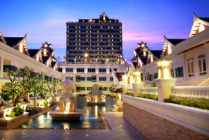 A lavish resort with traditional architectural elements is shown at dusk. The Grand Pacific Sovereign Resort & Spa features a central courtyard with illuminated fountains, surrounded by elegant buildings with intricate roof designs. A tall building is visible in the background against a purple sky.