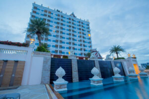 Property for sale by Auction Luxury hotel with a large pool and decorative sculptures on Ruamjit Road, Cha-Am, Petchaburi. The multi-story building features numerous balconies, lit windows, and is flanked by tall palm trees. The scene is set against a clear, slightly cloudy sky, creating a serene and inviting atmosphere. Presented by World of Auctions