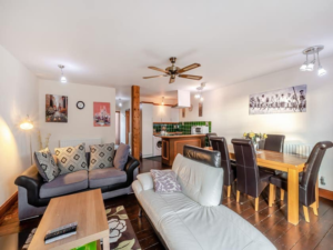 Property for sale by Auction A modern living and dining area with dark wooden flooring at The Old Drapery Apartments, 12 Main St, Haltwhistle. The room features a black and grey sofa, a white recliner, a wooden coffee table, and a dining table with black chairs. The kitchen has green tile backsplash. Artworks and a ceiling fan decorate the space. For sale by auction. Presented by World of Auctions