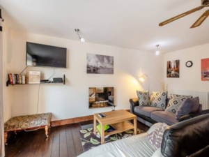 Property for sale by Auction A cozy living room with wooden flooring, featuring a wall-mounted TV above a shelf with books and decor in The Old Drapery Apartments, 12 Main St, Haltwhistle for sale by auction. Adjacent is a small bench, a comfy sofa with cushions, a rug, and a coffee table with a remote. Art prints adorn the walls, and a ceiling fan and lights provide illumination. Presented by World of Auctions