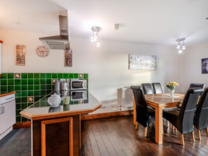 Property for sale by Auction A modern kitchen and dining area with wooden flooring at The Old Drapery Apartments, 12 Main St, Haltwhistle, for sale by auction. The kitchen features a green tiled backsplash, white cabinets, a microwave, and a kettle. An island counter separates it from the dining space, which has a wooden table with black leather chairs and a vase of flowers. Presented by World of Auctions