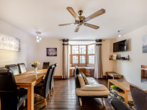 Property for sale by Auction A cozy living and dining room with wooden floors and modern furniture at The Old Drapery Apartments, 12 Main St, Haltwhistle—now for sale by auction. The dining area has a wooden table with six chairs, while the living area features a beige sofa, a wall-mounted TV, and a coffee table. A ceiling fan and large windows let in natural light. Presented by World of Auctions