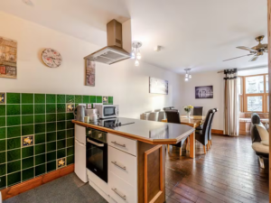 Property for sale by Auction A modern kitchen and dining area with wooden flooring at The Old Drapery Apartments, 12 Main St Haltwhistle, for sale by auction. The kitchen boasts a green tiled backsplash, white cabinets, and a central island with a stovetop and range hood. A wooden table set for six graces the dining area, complemented by artwork and a fan. Presented by World of Auctions