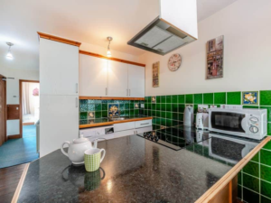 Property for sale by Auction A modern kitchen with green tiled backsplash and white cabinets in The Old Drapery Apartments at 12 Main St Haltwhistle, for sale by auction. The countertop features a teapot, a cup, and a kettle. Appliances such as a microwave, coffeemaker, and oven are visible. A clock and paintings adorn the well-lit space. Presented by World of Auctions