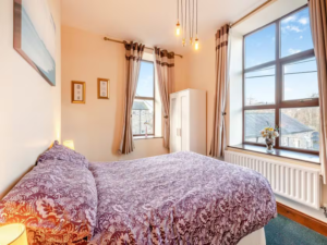 Property for sale by Auction A cozy bedroom at The Old Drapery Apartments, 12 Main St, Haltwhistle, featuring a double bed with a purple and white patterned bedspread. The room boasts two large windows with beige curtains, a white wardrobe, wall art, framed certificates, hanging lights, and a vase of flowers on the windowsill. This charming property is for sale by auction. Presented by World of Auctions