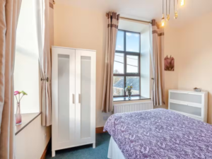 Property for sale by Auction A cozy bedroom in The Old Drapery Apartments at 12 Main St, Haltwhistle, features a large window with cream curtains, beige walls, and a green carpet. The bed is adorned with a purple patterned duvet, while the white wardrobe boasts frosted glass panels. Flower decorations enhance the ledge and dresser. For sale by auction. Presented by World of Auctions