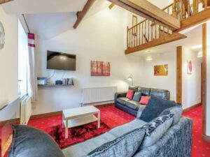 Property for sale by Auction A cozy living room at The Old Drapery Apartments, 12 Main St Haltwhistle, with a high ceiling and wooden beams. It features a gray and black sofa with decorative cushions, a white coffee table on a red rug, and a mounted flat-screen TV. The room also has a staircase and wooden railing leading to a loft area. For sale by auction. Presented by World of Auctions