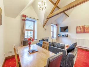 Property for sale by Auction A cozy living and dining area at The Old Drapery Apartments, 12 Main St, Haltwhistle. Featuring wooden beams on a high ceiling, it includes a wooden dining table, chairs, a black sofa, wall-mounted TV, and artwork. Red carpet, potted plants, and large windows with curtains complete the inviting atmosphere. For sale by auction. Presented by World of Auctions