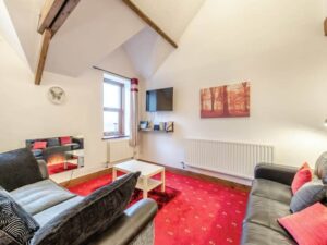 Property for sale by Auction A cozy living room with red carpet, two black leather sofas, and a white coffee table. The room features a wall-mounted flat-screen TV, a fireplace with a mirror above it, and a window with curtains. Ceiling beams are visible in this charming unit at The Old Drapery Apartments, 12 Main St Haltwhistle for sale by auction. Presented by World of Auctions