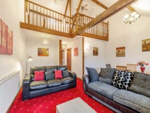 Property for sale by Auction A cozy living room at The Old Drapery Apartments, 12 Main St, Haltwhistle, features two sofas, a coffee table, and a vibrant red carpet. The ceiling is high with exposed wooden beams and a mezzanine level overlooks the space. Warm lighting and landscape artwork decorate the white walls, creating a welcoming atmosphere. For sale by auction. Presented by World of Auctions