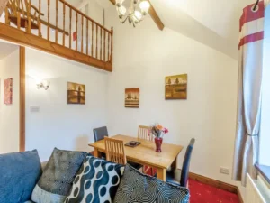 Property for sale by Auction A cozy dining area at The Old Drapery Apartments, 12 Main St, Haltwhistle, features a wooden dining table with four chairs, a decorative vase with flowers, and a couch with patterned pillows in the foreground. Artwork adorns the walls, and a mezzanine balcony is visible above. A chandelier hangs from the ceiling. For sale by auction. Presented by World of Auctions