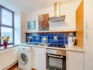 Property for sale by Auction A modern kitchen at The Old Drapery Apartments, 12 Main St, Haltwhistle, for sale by auction. Features a white countertop, electric stove and oven. There’s a blue-tiled backsplash with floral designs, two framed pictures above, and a washing machine by the sink. A white kettle and vase with flowers are on the counter. Large window in the background. Presented by World of Auctions