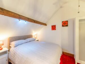 Property for sale by Auction A cozy bedroom in The Old Drapery Apartments, 12 Main St, Haltwhistle, features a double bed with a white comforter and two bedside tables with lamps. Above the bed is a wooden beam. The walls are adorned with two red rose paintings, and the room is illuminated with warm lighting. For sale by auction. Presented by World of Auctions