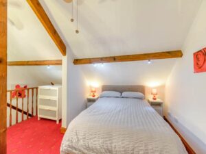 Property for sale by Auction A cozy attic bedroom at The Old Drapery Apartments, 12 Main St Haltwhistle, for sale by auction. The room features a double bed beneath a sloped ceiling with exposed wooden beams, flanked by nightstands with red lamps. A white dresser sits near the railing, and the room has red carpeting and soft, warm lighting. Presented by World of Auctions