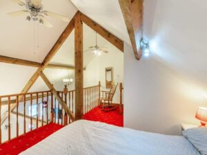 Property for sale by Auction A cozy bedroom loft at The Old Drapery Apartments, 12 Main St, Haltwhistle. Featuring wooden beams, a double bed with a white bedspread, red carpeted flooring, and a wooden rocking chair on the landing. Ceiling fans and a chandelier create a warm and inviting atmosphere. For sale by auction. Presented by World of Auctions