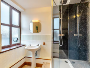 Property for sale by Auction A well-lit bathroom at The Old Drapery Apartments, 12 Main St Haltwhistle for sale by auction features a frosted window, white-tiled walls, and wood accents. Highlights include a sink with a small mirror above it, a round vanity mirror on the windowsill, and a glass-enclosed shower with dark, textured wall tiles. Presented by World of Auctions