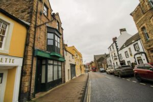 Property for sale by Auction A narrow street lined with historic buildings, including The Old Drapery Apartments at 12 Main St Haltwhistle for sale by auction. The exteriors, a mix of stone and brick in yellows, beiges, and whites, feature unique windows. Parked cars and signs wet from rain are visible under the overcast sky. Presented by World of Auctions