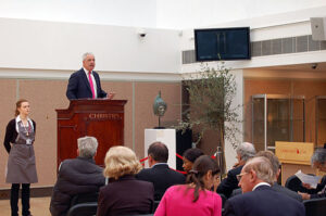 Property for sale by Auction An auctioneer stands behind a podium labeled "Christie's" speaking to a seated audience in an auction room. A screen displays auction details, and a sculpture on a pedestal is visible in the background. An assistant in an apron stands to the left of the podium. Presented by World of Auctions