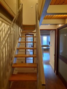 Property for sale by Auction A narrow wooden staircase with metal railings ascends to an upper floor. The area features wooden flooring and ceiling. In the background, there is a sunlit room with a large window and a table, accessible through a doorway. Presented by World of Auctions