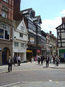 Property for sale by Auction A bustling street in a city with people walking and crossing. The street is lined with both modern and Tudor-style buildings with shops, including a branch of TSB bank and a café named "EAST." The sky is clear, indicating a bright day. Presented by World of Auctions