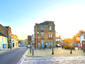 Property for sale by Auction A quaint town square with a historic brick building in the center. The sun sets, casting a warm glow over the scene. Cobblestone streets and a variety of small shops line the area, giving a charming and peaceful atmosphere. Nearby, The Old Drapery Apartments at 12 Main St Haltwhistle are for sale by auction. Presented by World of Auctions