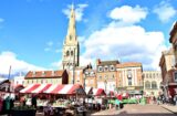 Property for sale by Auction A vibrant marketplace with stalls featuring red and white striped awnings, located in a city square. A prominent church tower rises in the background, surrounded by historic buildings under a bright blue sky with scattered clouds. People are browsing the market. Presented by World of Auctions