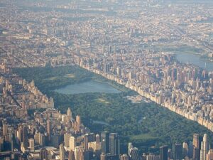 Property for sale by Auction Aerial view of Central Park in New York City, surrounded by tall buildings and skyscrapers. The park is lush and green, with visible bodies of water and tree-lined walking paths. The dense urban landscape extends outwards, with rivers visible in the background. Presented by World of Auctions
