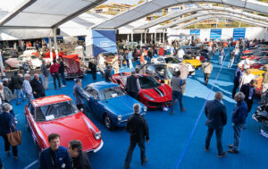 Property for sale by Auction A lively car show under a large white tent features numerous vintage and modern cars. People are casually walking around, inspecting the vehicles. The area is brightly lit with a blue carpeted floor, showcasing a variety of colorful cars parked neatly in rows. Presented by World of Auctions