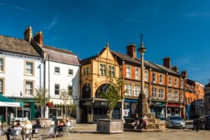 Property for sale by Auction A sunny day in a town square featuring colorful, historic buildings. People are sitting at outdoor tables under umbrellas near a pizza and fish bar, with more shops lining the street. A stone monument with a cross stands prominently in the center—prime residential development opportunity in Grantham. Presented by World of Auctions