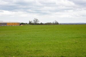 Property for sale by Auction A vast green field under a cloudy sky with a wooden and metal building on the left side. The landscape is mostly flat, with a few scattered leafless trees in the background and rolling hills visible in the distance. Presented by World of Auctions