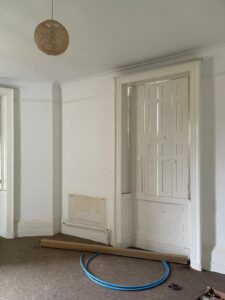 Property for sale by Auction An empty room with white walls and a high ceiling featuring a decorative light fixture. There are two tall, narrow windows and a white, panelled door. The floor is carpeted, and a blue hose and a cardboard tube are lying on the floor. Presented by World of Auctions