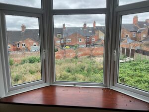 Property for sale by Auction A view through a bay window shows a backyard area overgrown with grass and weeds. Beyond the yard, a row of brick houses with chimneys lines the background beneath a cloudy sky. The window sill has some scattered debris. Presented by World of Auctions