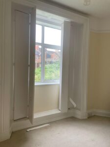 Property for sale by Auction A small room with beige walls and beige carpeting features a large window with white trim that opens up to show the view outside. The window is open and the shutters are partially open at different angles, offering a glimpse of houses and greenery in the background. Presented by World of Auctions