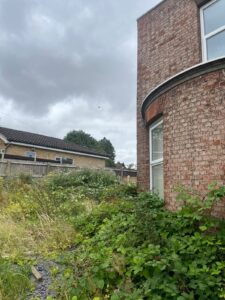 Property for sale by Auction A brick building with tall windows stands next to an overgrown garden filled with weeds and shrubs. In the background, a smaller house with a tiled roof is visible. The sky is overcast with dark clouds, indicating possible rain. Presented by World of Auctions