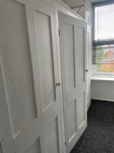 Property for sale by Auction A white wooden built-in wardrobe with paneled doors is situated in a corner of a room with white walls and dark carpet. The wardrobe is adjacent to a window with partially open blinds, allowing some natural light to enter and offering a view of houses and greenery outside. Presented by World of Auctions