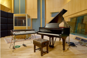 Property for sale by Auction A music studio with a grand piano, an electric keyboard on a stand, and a cushioned bench. The room has wooden floors and a modern design with acoustic panels on the walls. In the background, there is a glass vocal booth with a microphone inside. Presented by World of Auctions