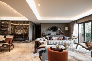 Property for sale by Auction A modern living room with marble flooring and a mix of contemporary and classic furniture. The space includes a stylish sofa with striped upholstery, a circular coffee table, various armchairs, decorative items, and large windows allowing natural light. Lush greenery is visible outside. Presented by World of Auctions