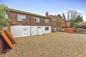 Property for sale by Auction A two-story brick building with a white lower facade has two white doors and multiple windows. The yard in front is covered with gravel, and there are stacks of wooden boards on the left side. Trees and foliage are visible in the background. Presented by World of Auctions